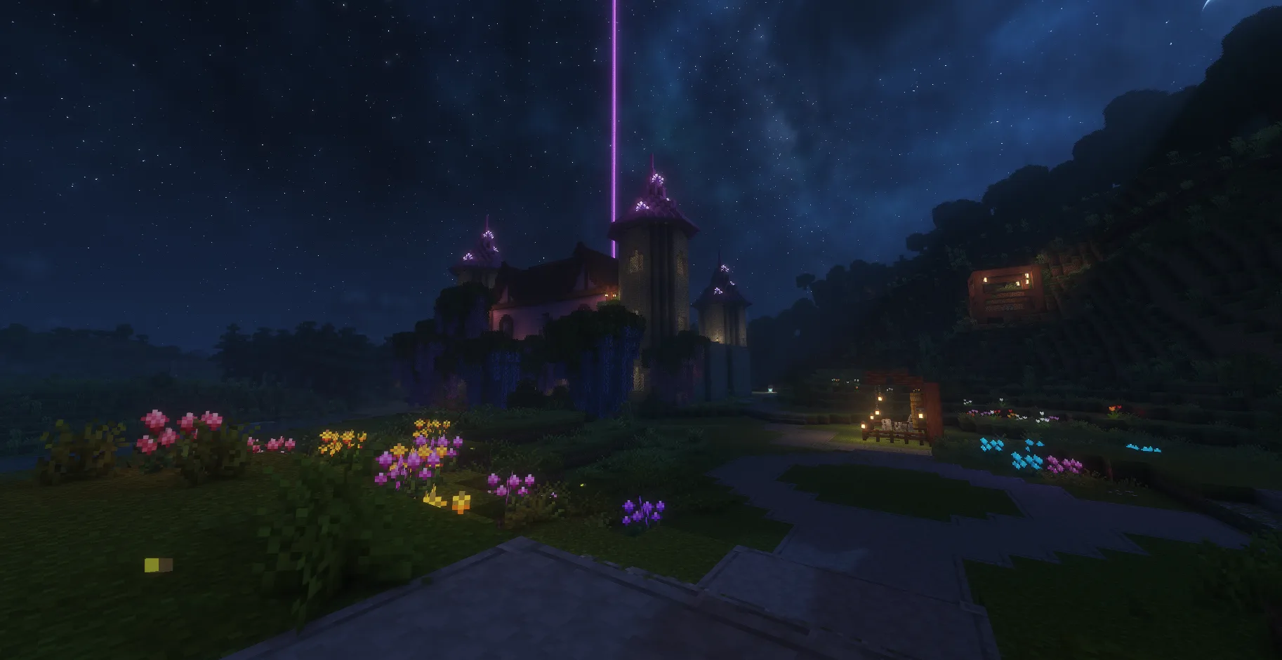 A purple-lit castle beneath a starry night, bordered by bright flower beds and a small lantern-lit seating area.