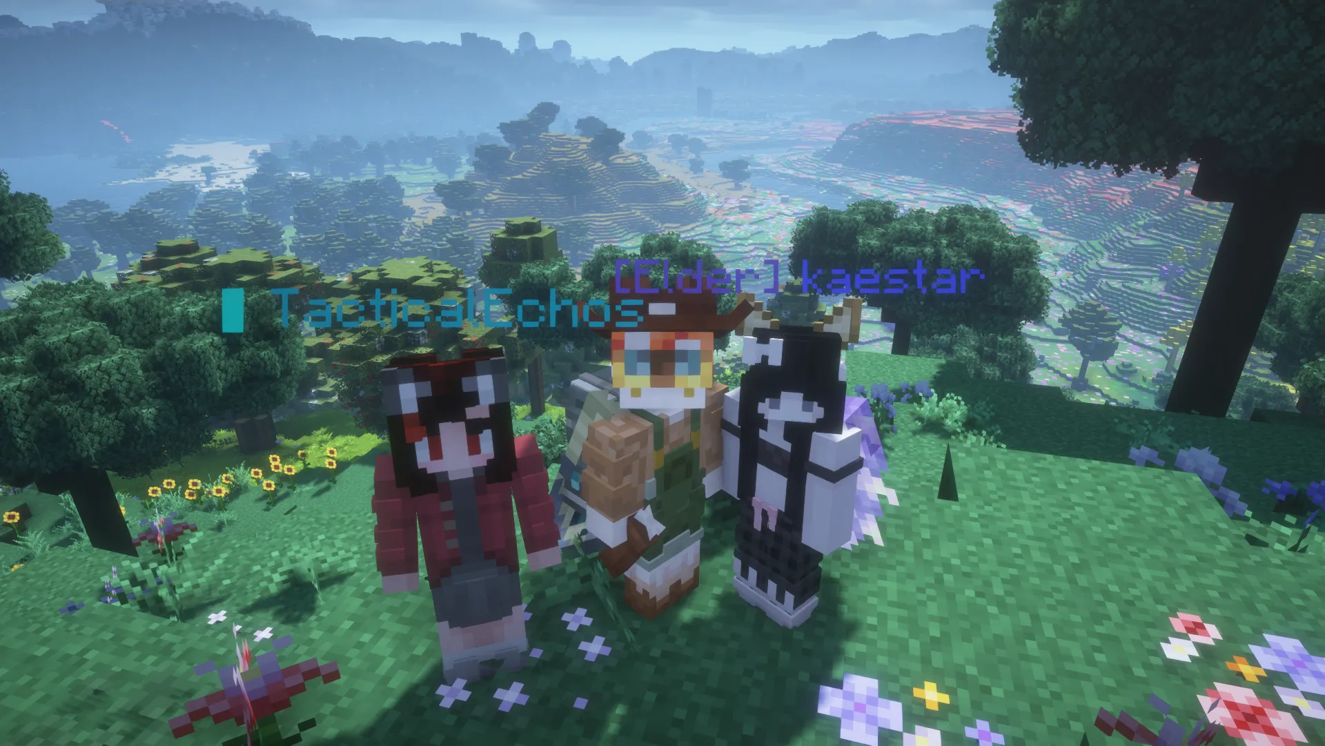 TacticalEchos, Omayor, and kaestar pose together on a flowered hillside above a sunlit forest valley.