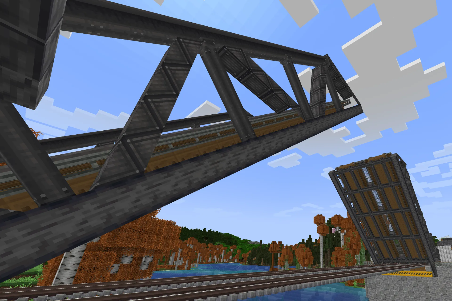 A dark iron truss railway bridge crosses above tracks and water, framed against an autumn forest and blue sky.