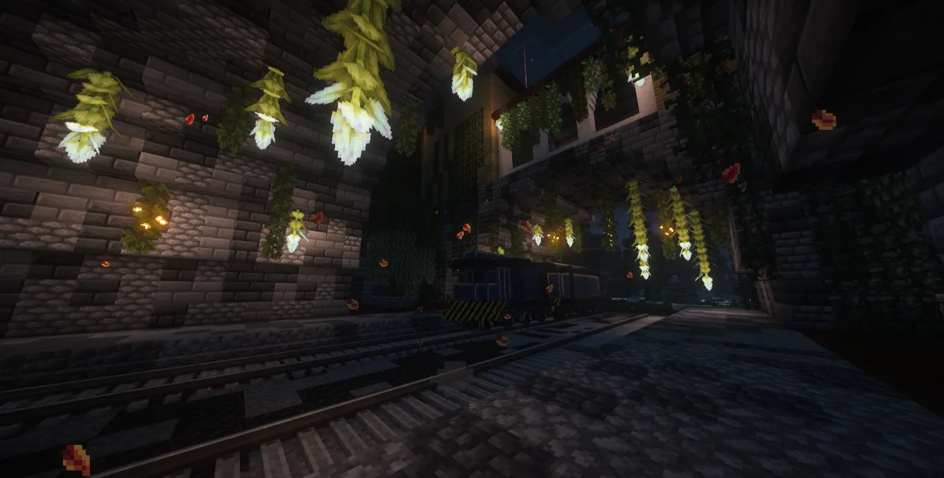 A dark brick railway hall lit by hanging flowers and lanterns, with a freight train waiting on twin tracks.