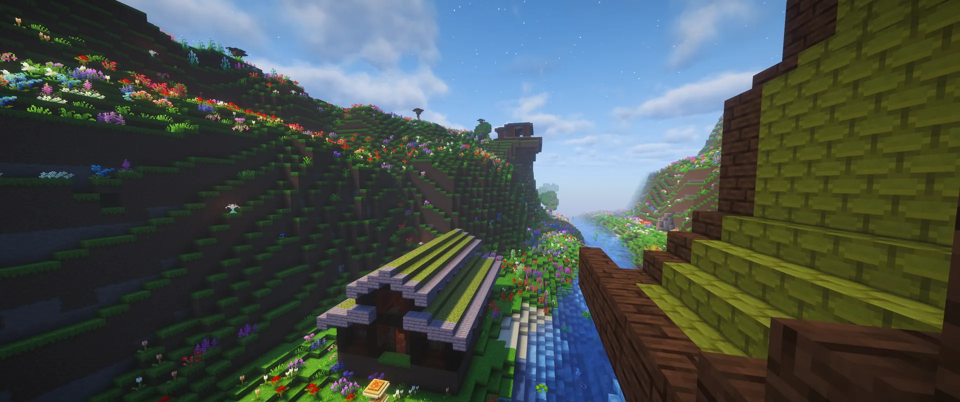 Green-roofed buildings beside a narrow river in a flower-covered valley, seen from a neighboring roof under a bright blue sky.