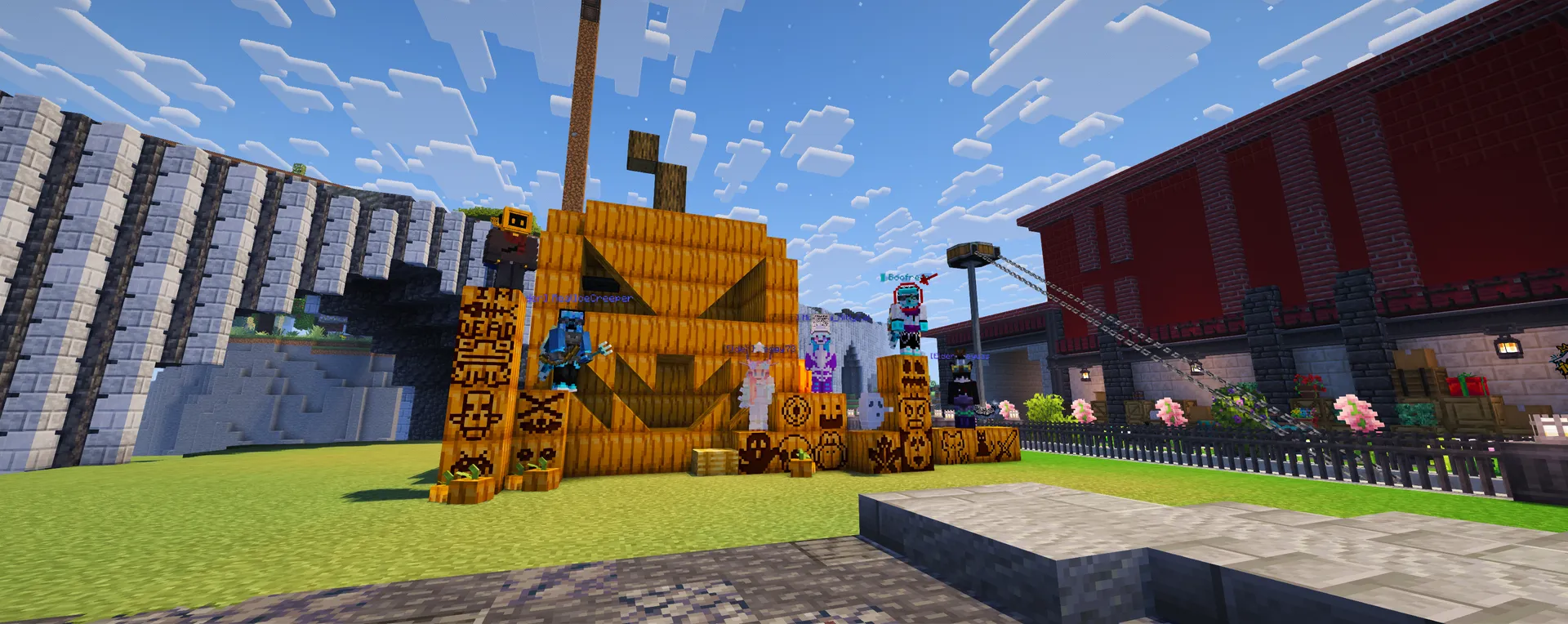Players pose among stacks of carved pumpkins beside a giant jack-o’-lantern, with a red building and flower beds behind them.